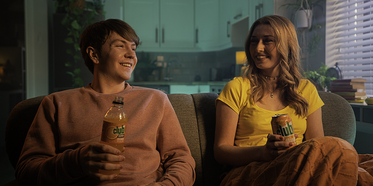 Image of two people sat on a sofa - one is holding a bottle of Club Orange and the other with a can of Club Orange.