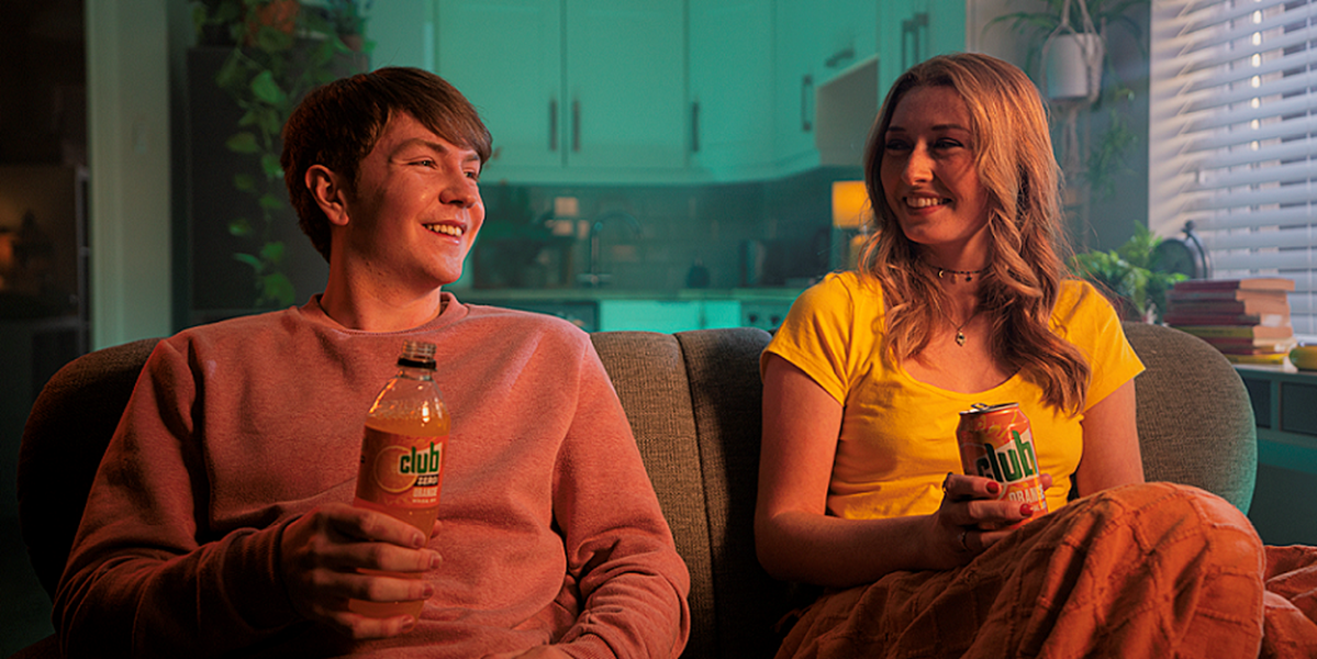 Image of two people sat on a sofa - one is holding a bottle of Club Orange and the other with a can of Club Orange.